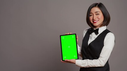 Woman concierge presenting greenscreen display on tablet, showing chromakey template on screen. Receptionist with bow and uniform holding device with blank copyspace layout. Camera B.