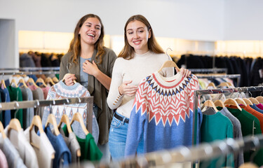 Cheerful female friends in casual outfit admiring pullovers with a pattern in a showroom
