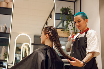 Beautiful hair stylist with tattooed hands applying spray on woman wet hair in beauty salon