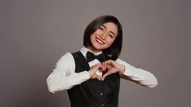 Hotel administrator shows heart shape symbol on camera, expressing romantic gesture and being flirty. Asian receptionist presenting true honest feelings, does romance sign in studio. Camera B.