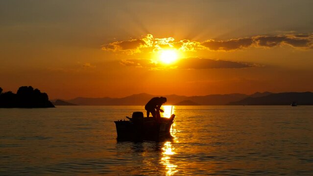 Man floating on vessel by evening coastline. A man silhouette floating on wooden vessel on lake against orange dusk sky in summer. A concept of swimming hobby.
