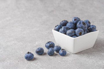 Fresh blueberries in a small square white bowl with scattering of berries on gray concrete background with copy space for text. Organic berries, healthy food, wild berries