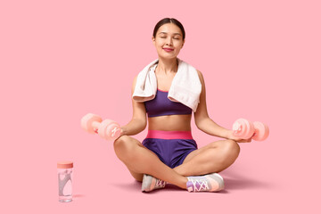 Young woman in sportswear and with dumbbells meditating against pink background
