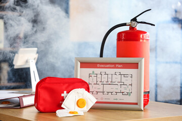 Evacuation plan with masks, first aid kit and fire extinguisher on table in burning office building