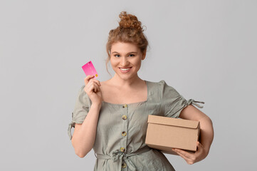 Beautiful young woman with credit card and cardboard box on grey background