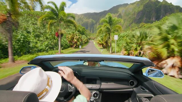 Happy girl in blue convertible car enjoying summer vacation, waiving hand out of the car and showing shaka hawaiian sign. Woman travels by tropical island with tropical exotic mountain landscape Oahu