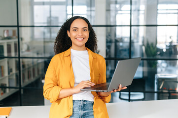 Successful confident beautiful hispanic business woman in stylish clothes, business coach, manager, standing in a modern office, holding an open laptop, looking and smiling at camera