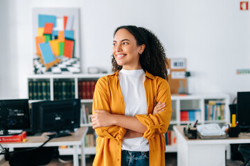Positive successful confident beautiful hispanic curly haired business woman in orange shirt, business coach, manager, standing in a modern office with arms crossed, looking away and smiling