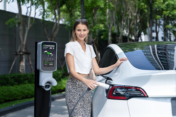 Young woman travel with EV electric car charging in green sustainable city outdoor garden in...
