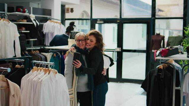 Senior woman and her daughter take photos in department store, enjoying day off shopping for clothes. Smiling customers have fun with selfies in a fashion showroom, commercial activity. Camera B.