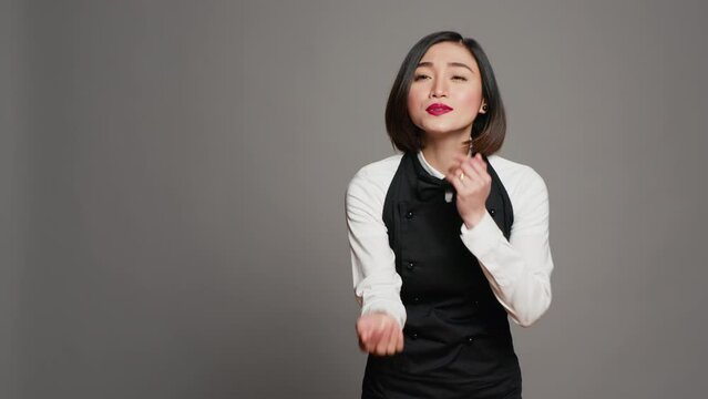 Cute flirty waitress blowing air kisses in front of camera, acting romantic with sweet gesture. Asian restaurant employee posing with a kissy facial expression, catering industry. Camera A.