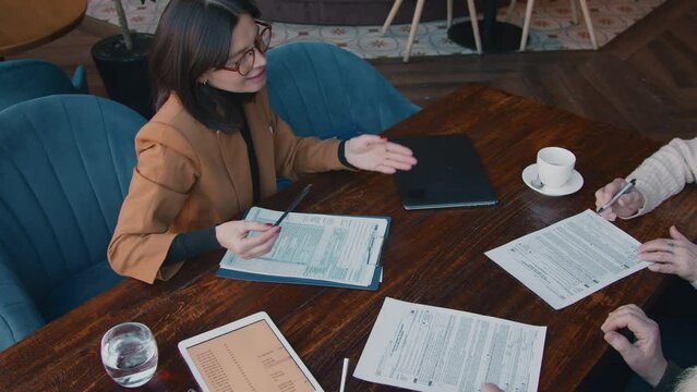 Medium top shot of hands of senior husband and wife sitting in cafe with financial consultant, signing insurance policy and legal documents, giving them back, woman smiling and thanking clients