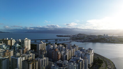 Bela imagem aérea das pontes de acesso a Florianópolis com prédios urbanos ao entardecer com o mar ao fundo.