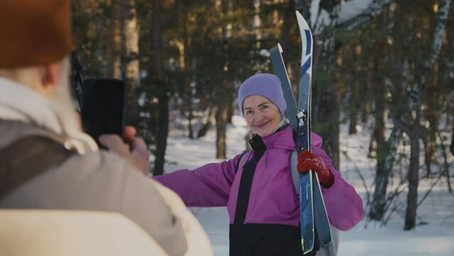 Medium over shoulder shot of senior man standing in winter forest and taking photos on smartphone of cheerful Caucasian woman, posing with skis and poles, smiling