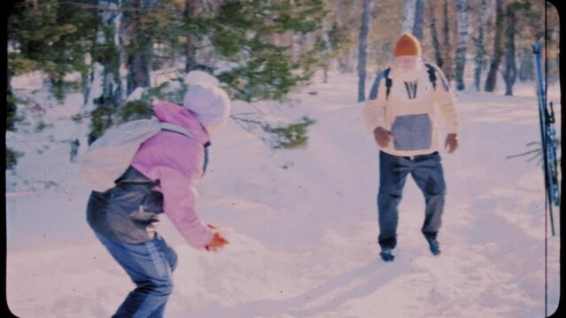 Full shot with grainy scratched retro film effect of elderly Caucasian husband and wife having playful snowball fight in winter forest, while resting during ski hike on sunny day