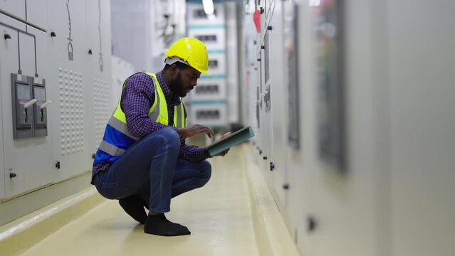 African man engineer in safety uniform working on digital tablet at factory server electric control panel. Industrial technician worker maintenance checking power system at manufacturing plant room.