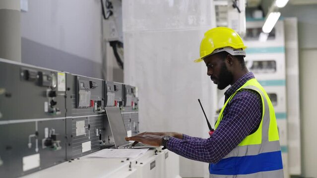 African man engineer in safety uniform working on laptop at factory server electric control panel room. Industrial technician worker maintenance checking power system at manufacturing plant room.