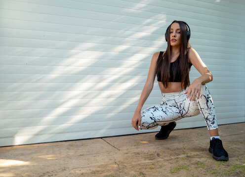 Sports fashion style photo, girl in modern leggings and headphones posing by the garage, kneeling. Showing confidence 
