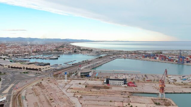Elevated drone shot capturing the sprawling cityscape alongside a busy commercial cargo shipping port with containers and cranes. The atmosphere is dynamic, with a clear blue sky. Valencia, Spain 