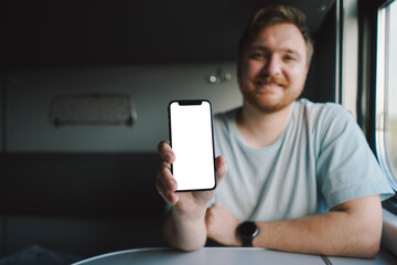 A man with a beard and mustache in a blue t-shirt is using a smartphone while traveling by Railway train, sitting in the train and looking out the window.