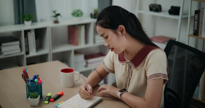 Footage high angle view, Selective focus,Happy young asian woman sitting at desk while thinking idea holding pen making notes in diary at home, creative thoughts to journaling