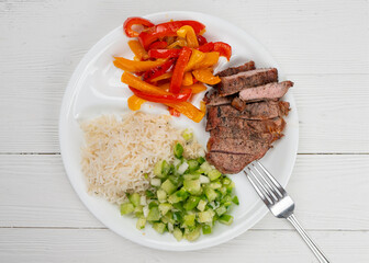 Meal. Fajita, sweet bell peppers, onions, green salsa tomatillo, beef ribeye steak grilled on charcoal and Basmati rice. Cooked meat and vegetables. Healthy dinner. Plate on white natural wooden table