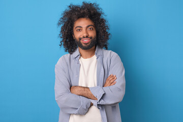 Young handsome successful Arabian man crosses arms in front of chest demonstrating confidence in himself and presence of ambition to achieve career goals stands on plain blue background.