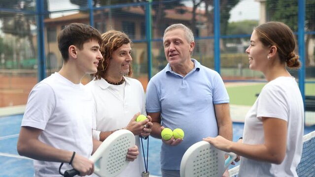 Four positive tennis players of different ages discussing preparing for padel game on tennis cort outdoors