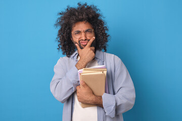 Young funny curly Arabian man student holds notebooks and squeezes face with hand showing dissatisfied grimace because of difficulty of exam or bad educational program stands on plain blue background.