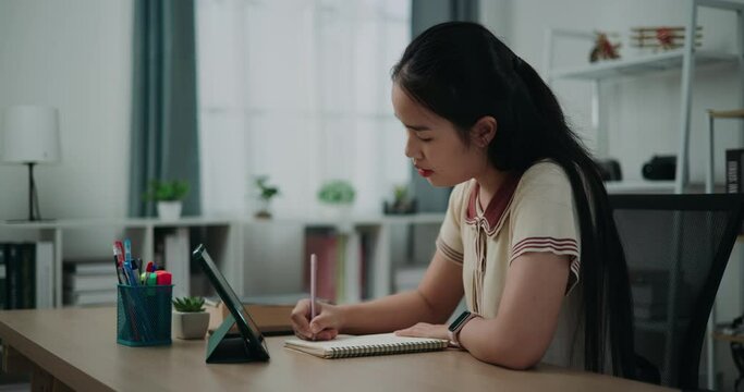 Footage dolly shot, Happy young asian woman sitting at desk writing notes while working on tablet for online learning at home, creative thoughts to journaling, idea and inspiration