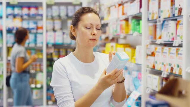 In pharmacy, woman carefully examines package of cream for problem skin. Client reads composition of therapeutic cosmetics on product