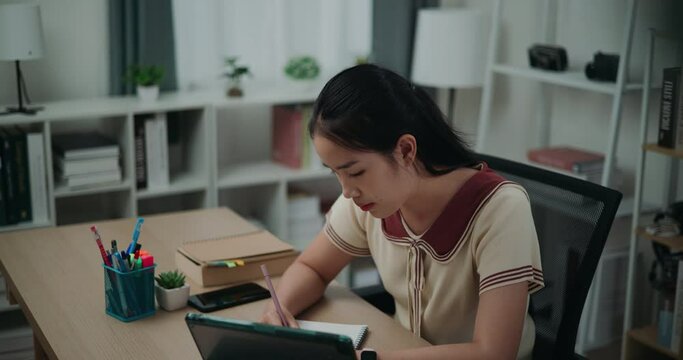 Footage dolly and high angle view shot, Happy young asian woman sitting at desk writing notes while working on tablet for online learning at home, creative thoughts to journaling, idea and inspiration