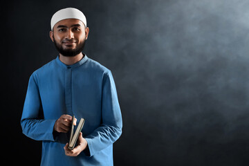 Portrait of handsome young asian muslim man with beard posing, holding holy book quran and smiling on dark background - 739687532