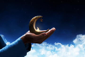 Hands of young asian muslim man holding shiny golden crescent moon with star lantern at blue night...