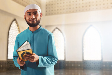 Portrait of handsome young asian muslim man with beard posing,  holding holy book quran and smiling in the mosque window arch - 739689323