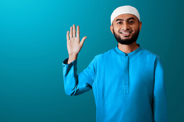 Portrait of handsome young asian muslim man with beard saying hello,  waving hand gesture and smiling isolated on blue studio background - 739690947