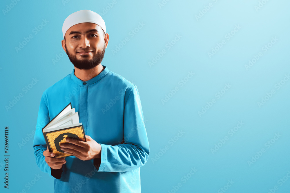 Wall mural portrait of handsome young asian muslim man with beard posing, holding holy book quran and smiling i