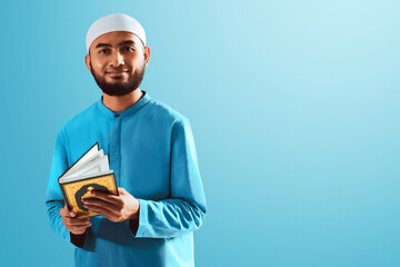 Portrait of handsome young asian muslim man with beard posing,  holding holy book quran and smiling isolated on blue studio background - 739691116