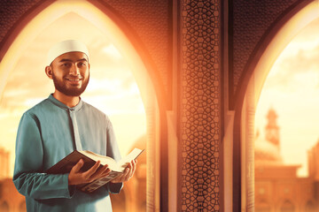 Portrait of handsome young asian muslim man with beard holding holy book quran and smiling in the mosque door arch - 739691394