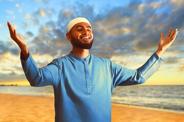 Portrait of handsome young asian muslim man with beard smiling, thankful and enjoying beautiful sunset on sea beach - 739691543