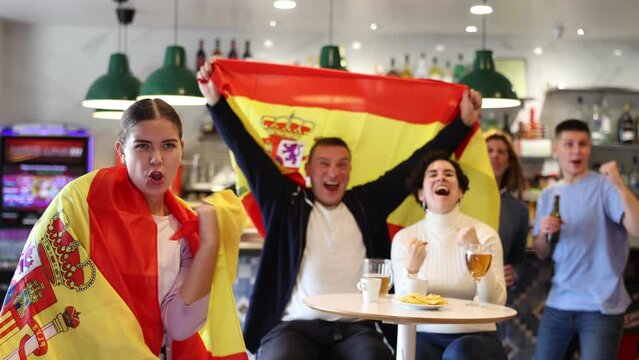 Company of emotive young adult sports fans supporting Spanish football team with state flag while resting in bar with beer