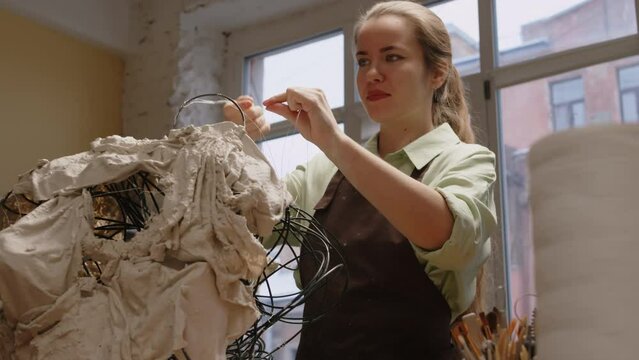 Tilt up low angle shot of concentrated young Caucasian female sculptor in apron bending armature wire to make shape, creating new piece of art with use of plaster cloths