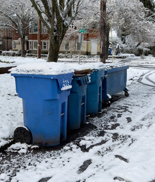 Blue plastic trash bins and a dumpster for household recyclable waste lined up curbside on a snow covered residential, neighborhood street 