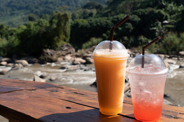 Two refreshing iced beverages of cantaloupe smoothie and strawberry soda on a wooden table with a serene river from waterfall and greenery in the nature background