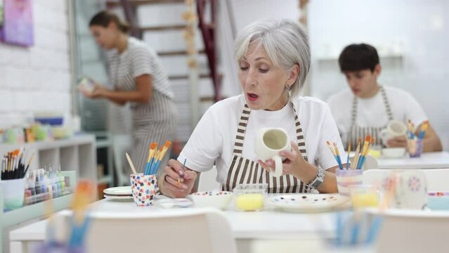 Interested elderly woman attending pottery class, enthusiastically painting on ceramic cup, creating unique custom design. Creative hobby concept