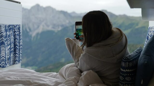 Young woman hold mug of coffee and taking pictures with smartphone of Dolomites from bed on travel van