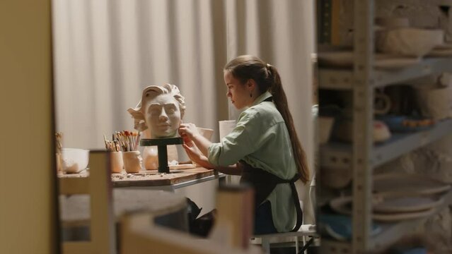 Side view medium shot of young Caucasian female sculptor making clay bust of young beautiful woman while working at desk in small creative workshopSide view medium shot of young Caucasian female sculp