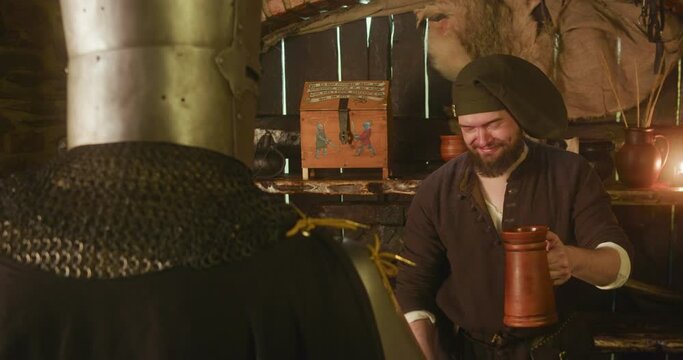 Servant gives drink with straw to knight in tavern. Happy dwarf innkeeper treats alcohol for lord in armour at bar counter. Adventurer visits old inn