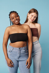 Vertical medium long studio portrait of two young ethnically diverse women with no makeup wearing bandeau tops and jeans posing for camera