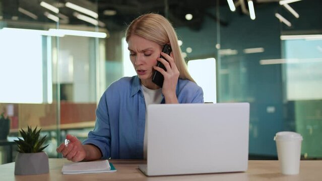 Attentive caucasian lady talking by mobile phone and making remarks in notebook by working table with laptop. Concentrated freelance copywriter getting edits from client and clarifying demands.